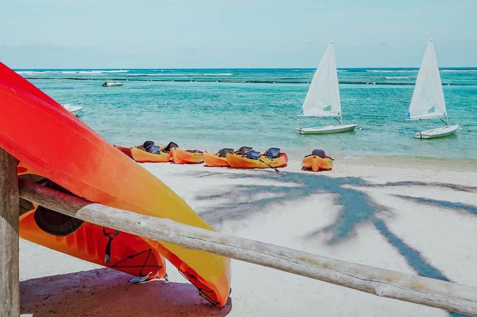Beach scene with colorful kayaks lined up on sand and white sailboats on turquoise water