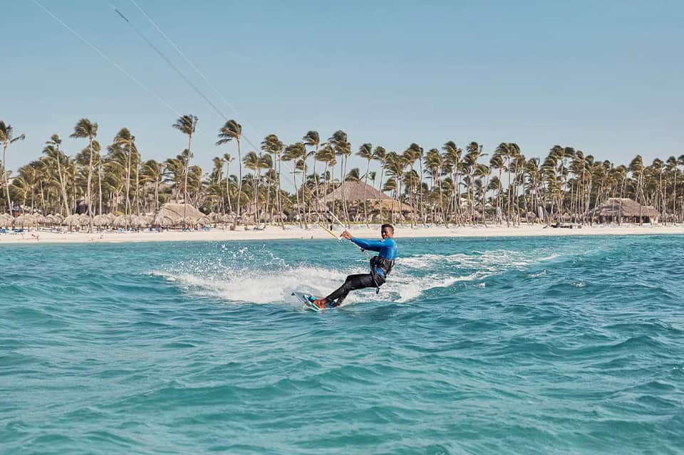 Kitesurfer in blue wetsuit riding waves with palm tree-lined beach and resort buildings in background