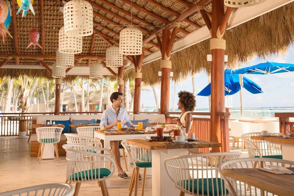 Beachfront restaurant with thatched roof, wooden beams, and couple dining with ocean view