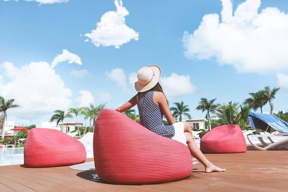Child in polka dot dress and straw hat sitting on pink bean bag by pool with palm trees