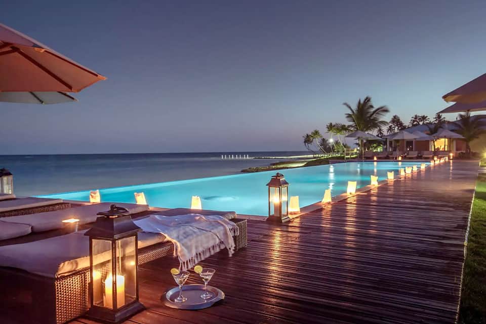 Illuminated poolside deck at dusk with lounge chairs, lanterns, cocktails, and ocean view
