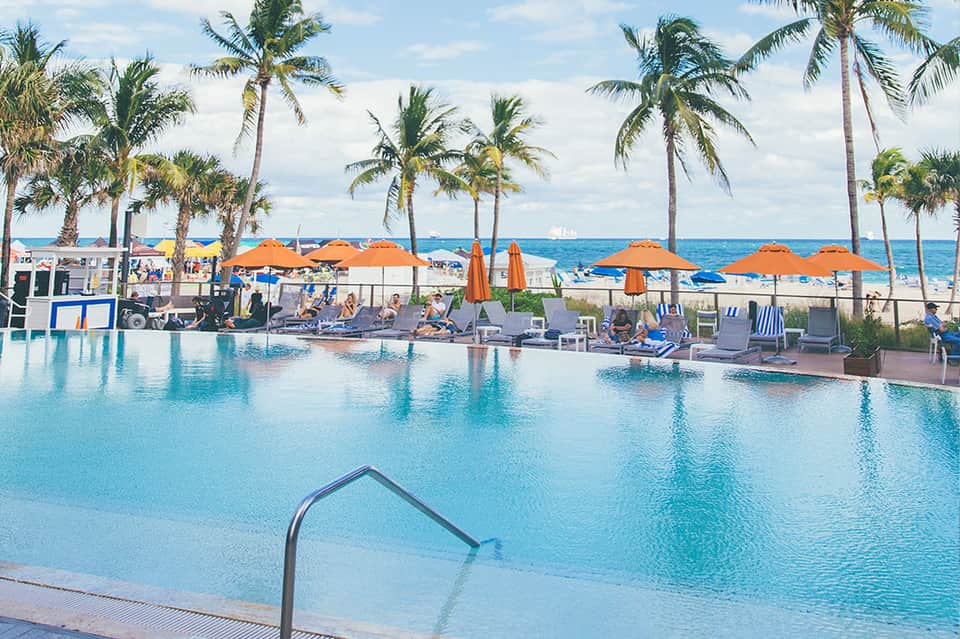 Resort infinity pool with palm trees, orange umbrellas, and beach access on sunny day