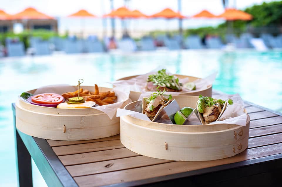 Bamboo steamer baskets with gourmet food and fries on a wooden deck overlooking a resort pool