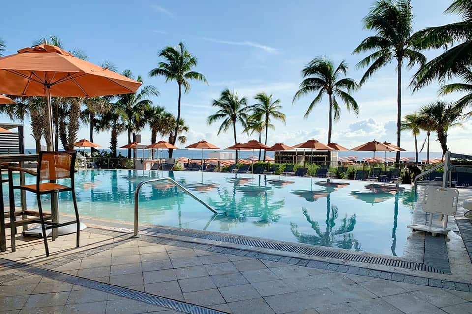 Resort pool area with turquoise water, orange umbrellas, palm trees, and lounge chairs on a sunny day