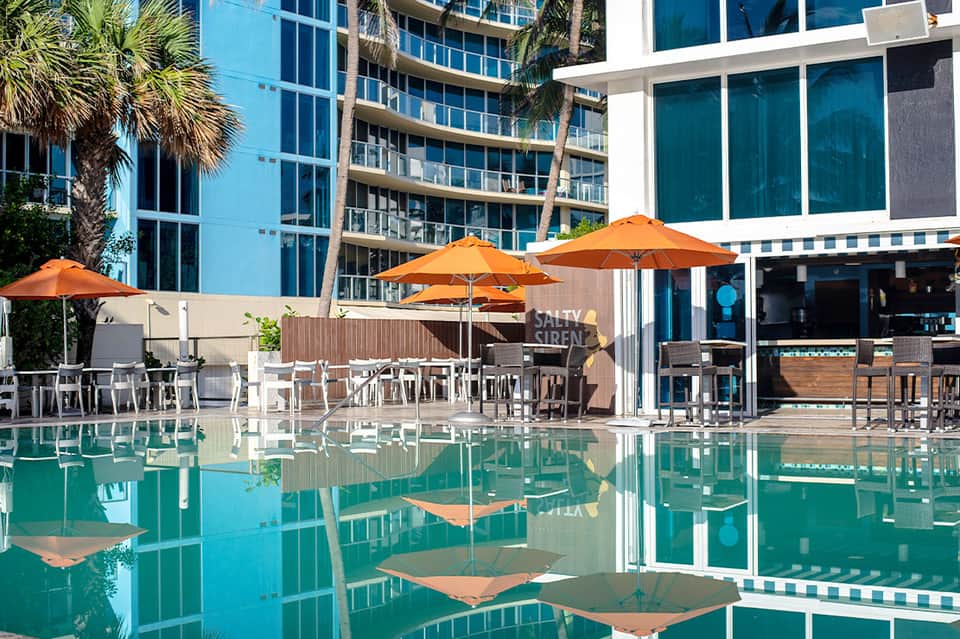 Resort pool deck with orange umbrellas, modern hotel building, bar area, and reflecting water