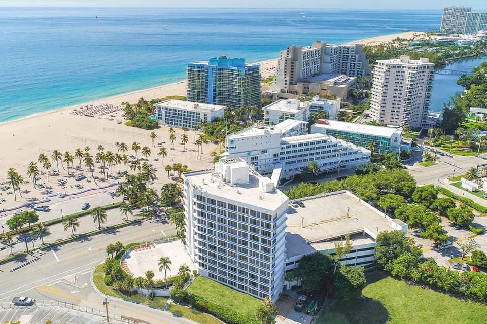 Aerial view of beachfront resort buildings with white sand beach, turquoise ocean, and surrounding high-rise hotels