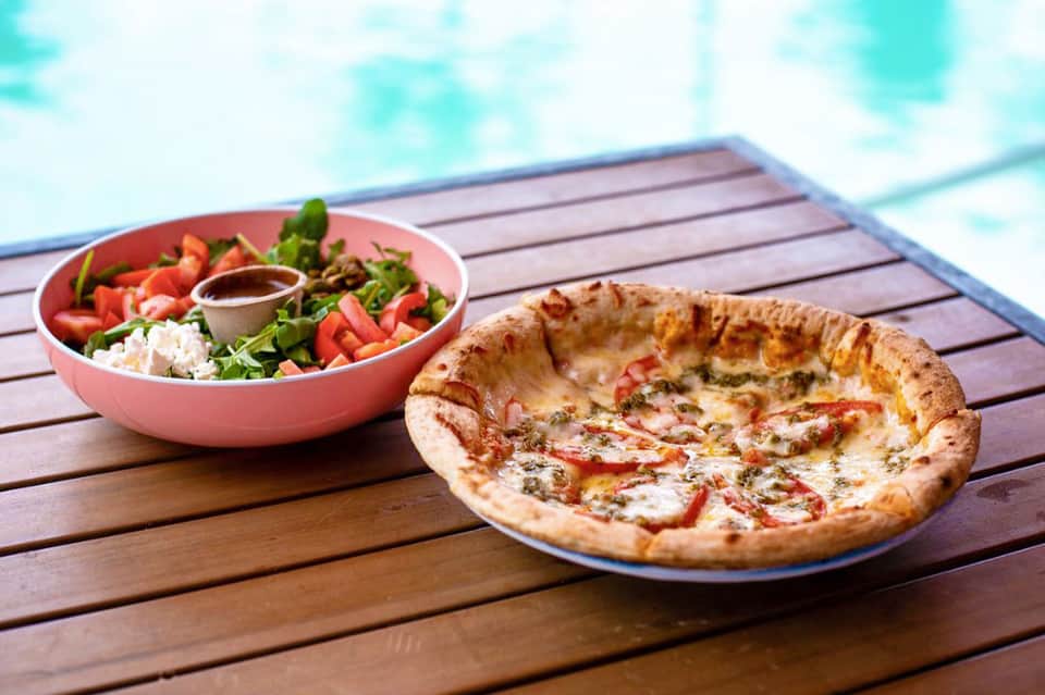Wood table with fresh salad bowl and baked pizza overlooking pool area