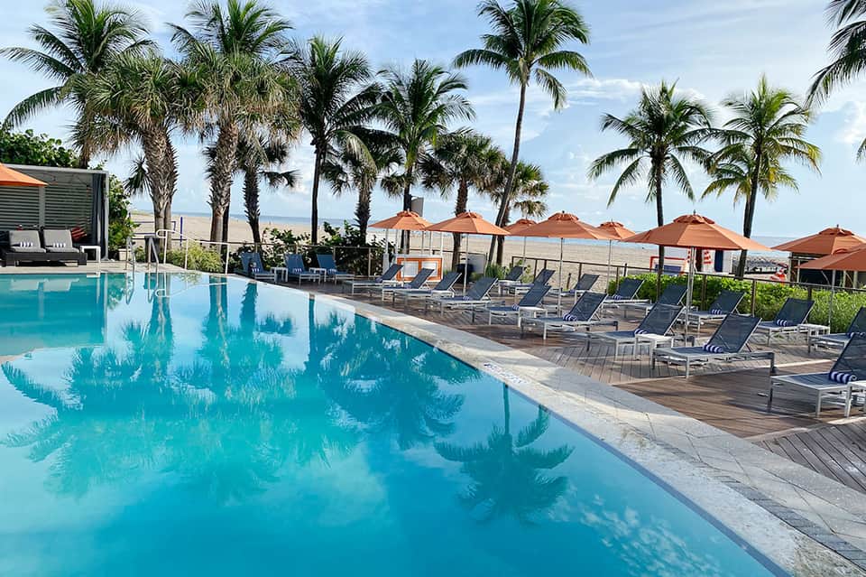 Luxury beachfront resort pool with palm trees, loungers, and orange umbrellas overlooking the sand and ocean