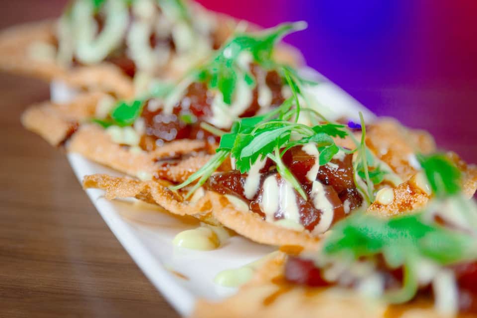 Gourmet appetizers on white plate featuring crispy flatbread with toppings and garnish against purple background