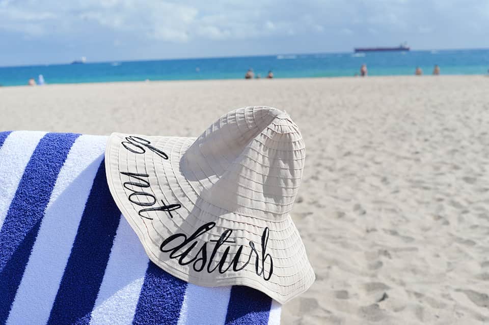 White sun hat with text resting on blue striped beach towel overlooking sandy beach