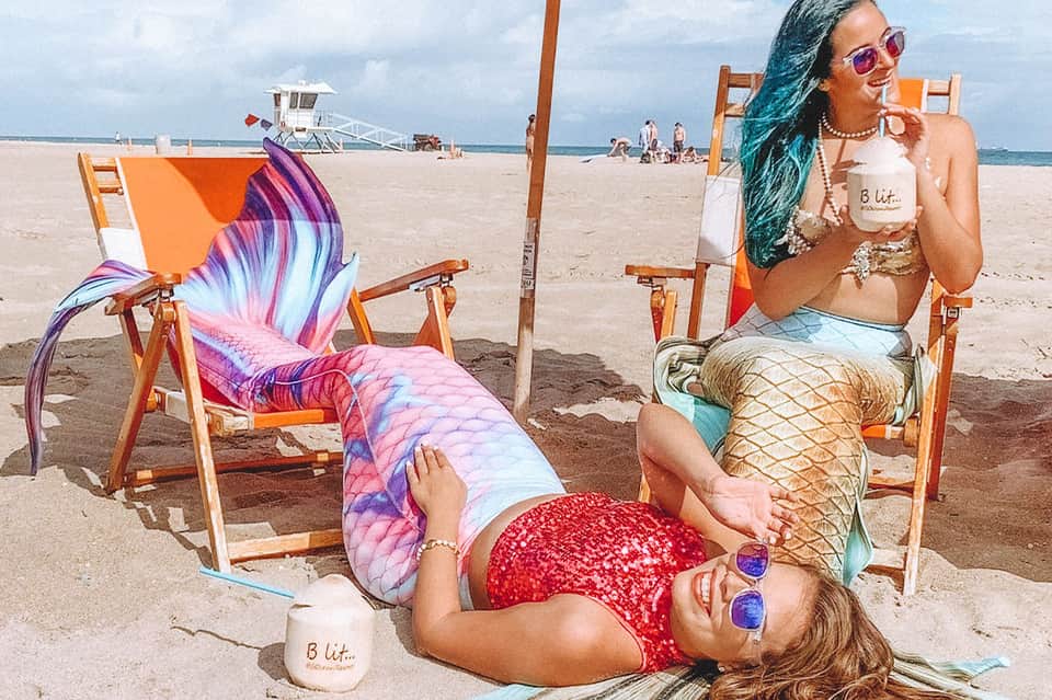 Two women in mermaid tails lounging on beach chairs with ocean and lifeguard tower behind