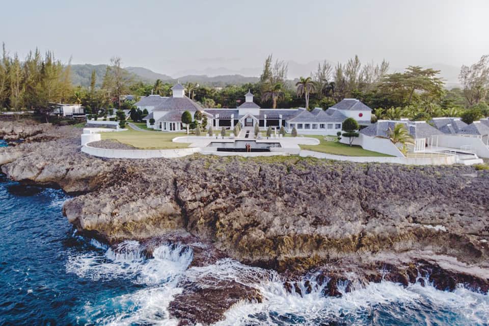 Luxury white seaside mansion on rocky coastal bluff with manicured lawns and mountain backdrop