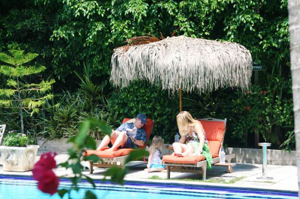 Family relaxing under thatched umbrella by pool with lush green landscaping
