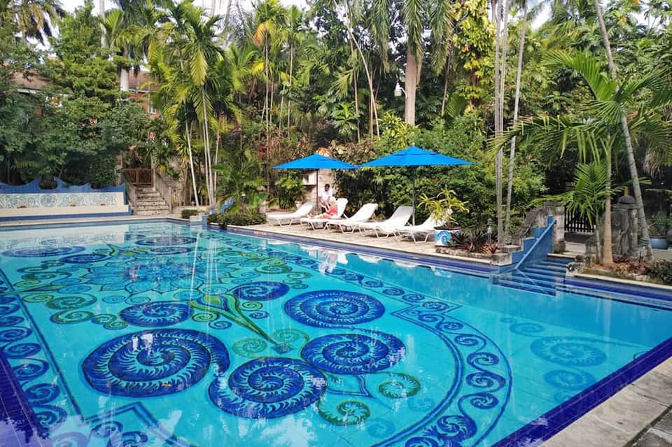Resort pool with decorative tile patterns, blue umbrellas, and lush tropical palms