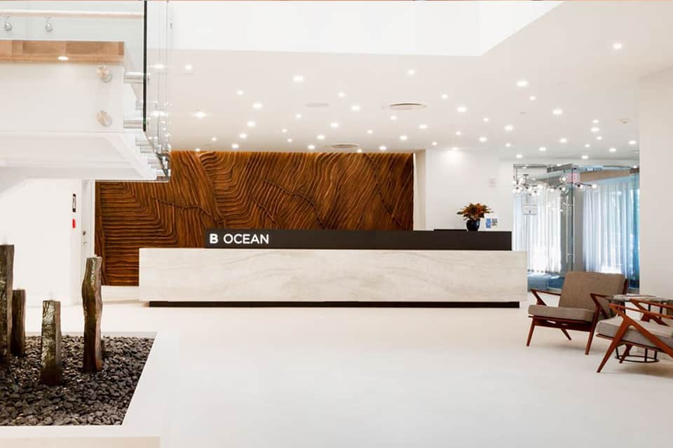 Contemporary hotel lobby with geometric wood accent wall, reception desk labeled 'B OCEAN', and minimalist design