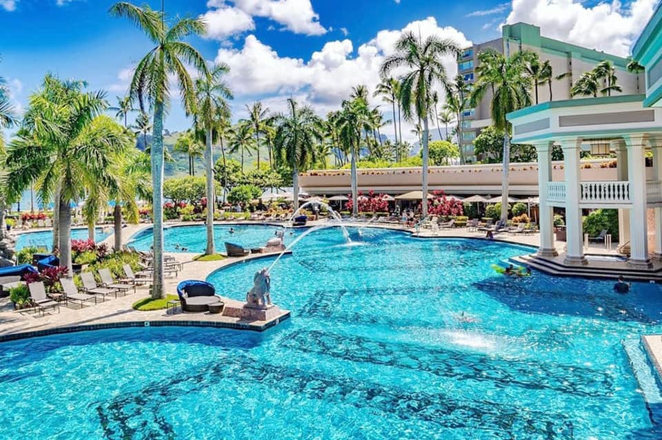 Resort pool complex with crystal-clear water, palm trees, white columns, and multi-level swimming areas
