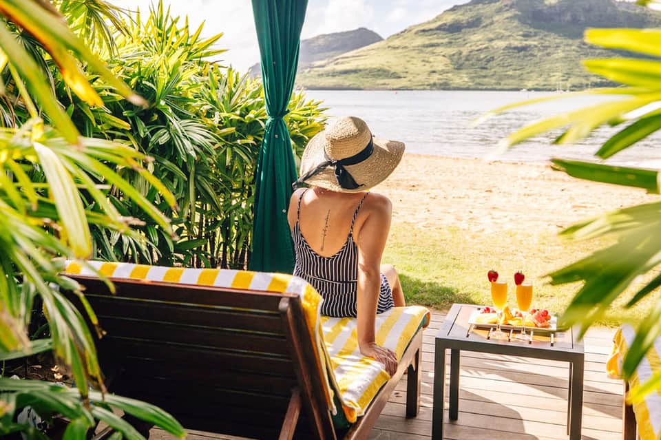 Woman in striped swimsuit and straw hat relaxing on a beach deck with tropical drinks and mountain views