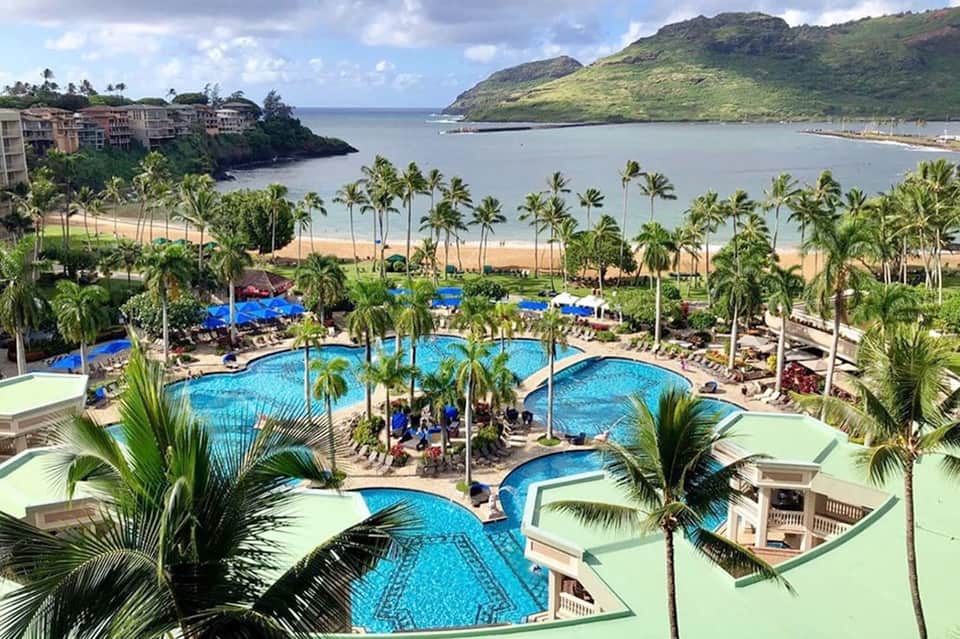 Beachfront resort with multiple curved pools, palm trees, and ocean view with green hills in background