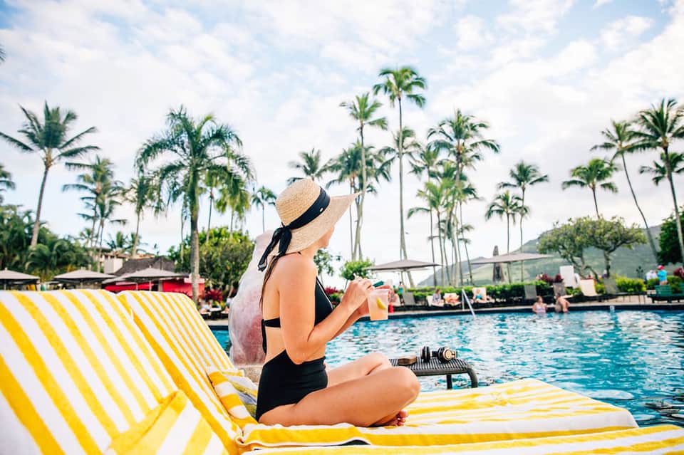 Woman in straw hat and black bikini sitting on yellow and white striped lounge chair holding drink by pool with palm trees