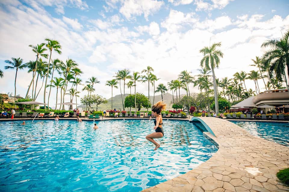 Woman jumping into resort pool surrounded by palm trees and lounge chairs with mountain backdrop