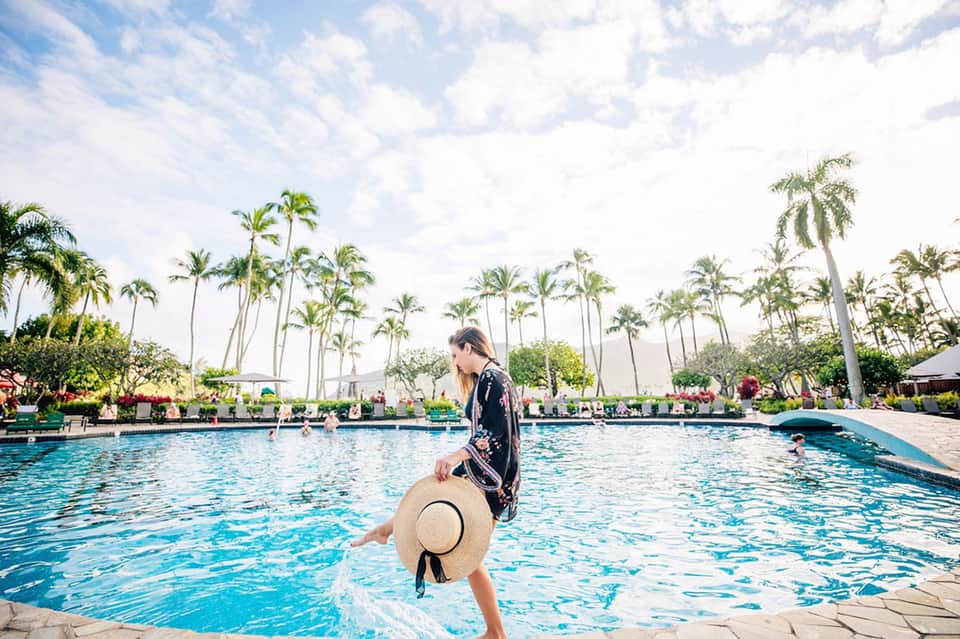 Woman in sunhat wading into large resort pool surrounded by palm trees and poolside loungers