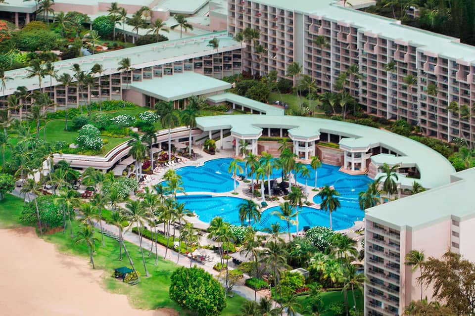 Aerial view of a large resort with curved pools, pink buildings, and palm trees
