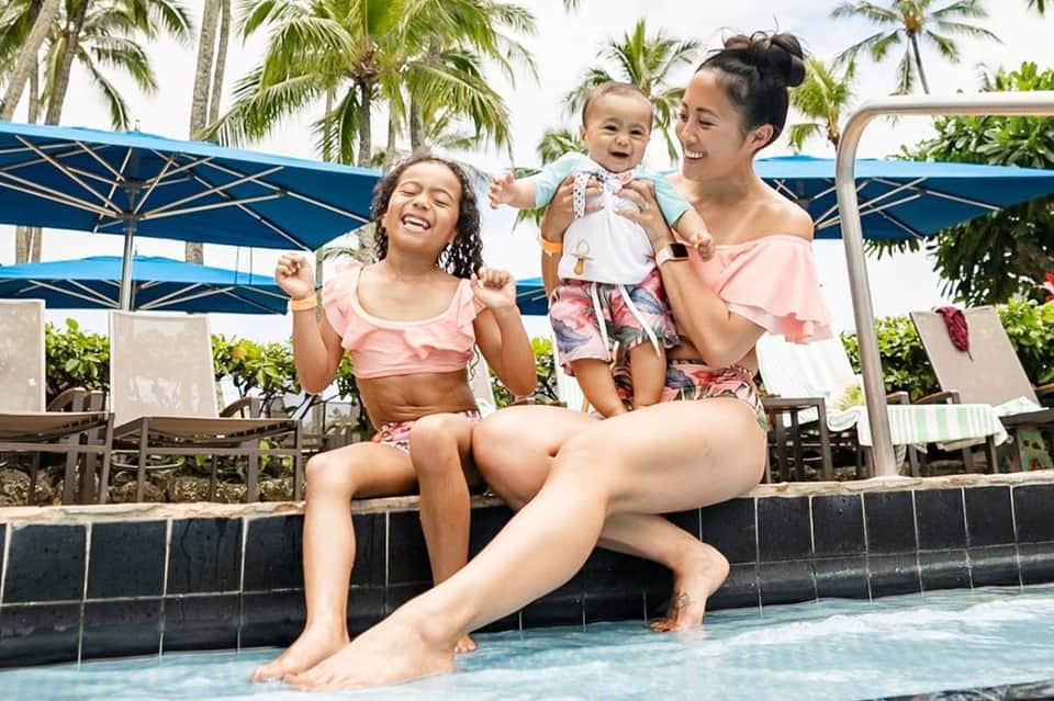 Family relaxing poolside with palm trees, blue umbrellas, and tropical surroundings