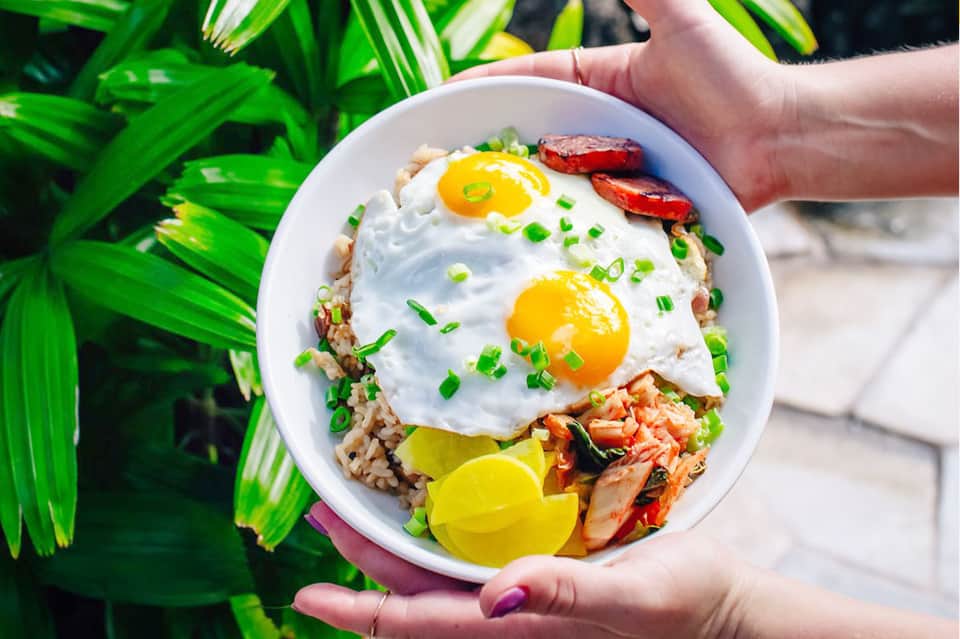 Hands holding bowl of rice with fried eggs, bacon, and lime garnish by tropical plants