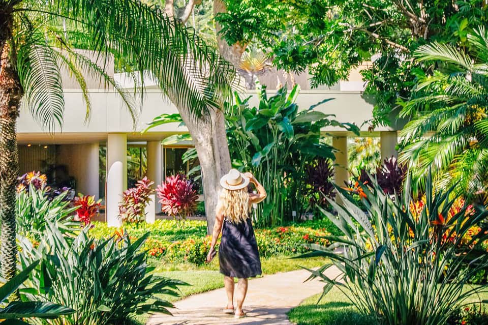 Woman in black dress and white hat walking through lush tropical garden courtyard