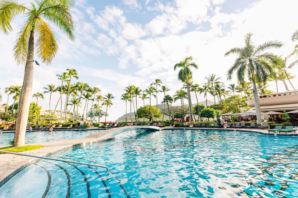 Large resort swimming pool surrounded by palm trees with mountains in tropical landscape background
