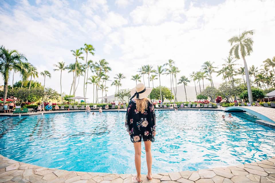 Woman standing poolside wearing a floral cover-up and sun hat at a tropical resort with palm trees