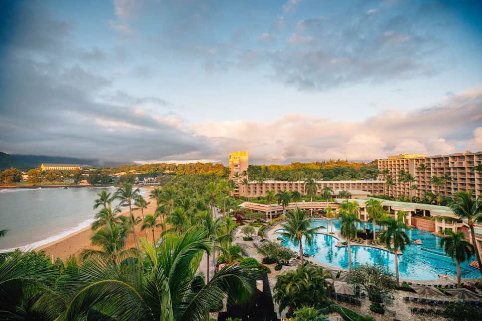 Beachfront resort aerial view with pool, palm trees, sand beach, and multi-story hotel buildings