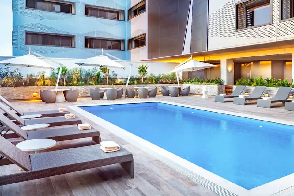 Resort courtyard with rectangular pool, lounge chairs, white umbrellas, and modern building facade