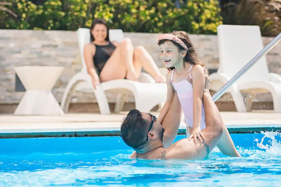Father and daughter playing together in resort swimming pool with lounge chairs