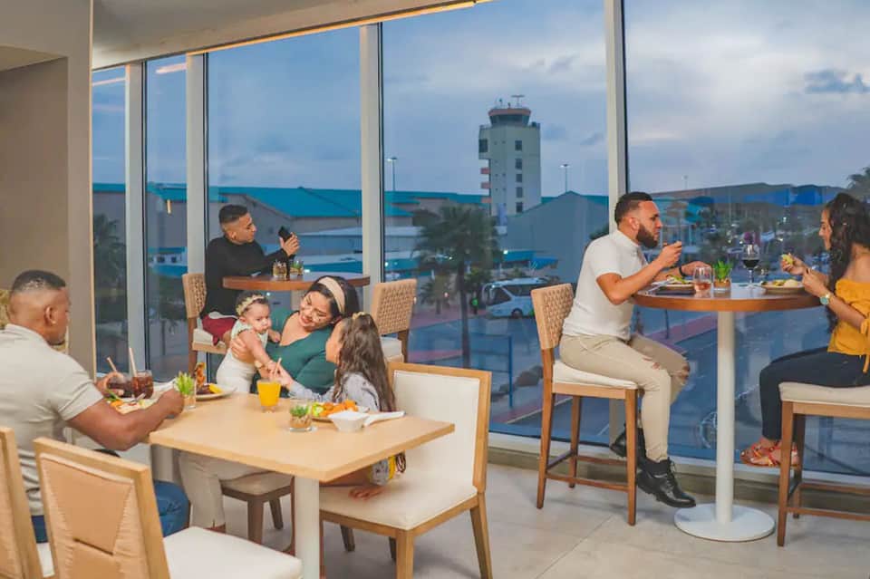 Families dining at tables near floor-to-ceiling windows overlooking a coastal harbor and tower with blue water views