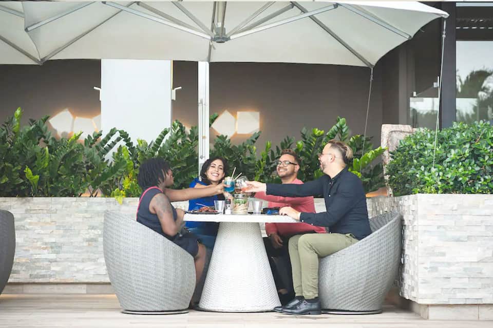 Four people toasting drinks at a round table on a modern terrace with white umbrellas and greenery