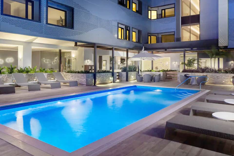 Modern hotel courtyard with illuminated blue pool, lounge chairs, and multi-story building at dusk