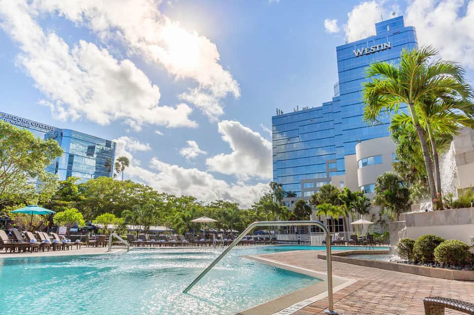 Modern resort pool area with turquoise water, palm trees, lounge chairs with umbrellas, and glass-fronted hotel building