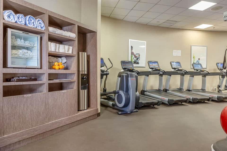 Hotel fitness center with treadmills and equipment, wooden storage unit with towels and amenities