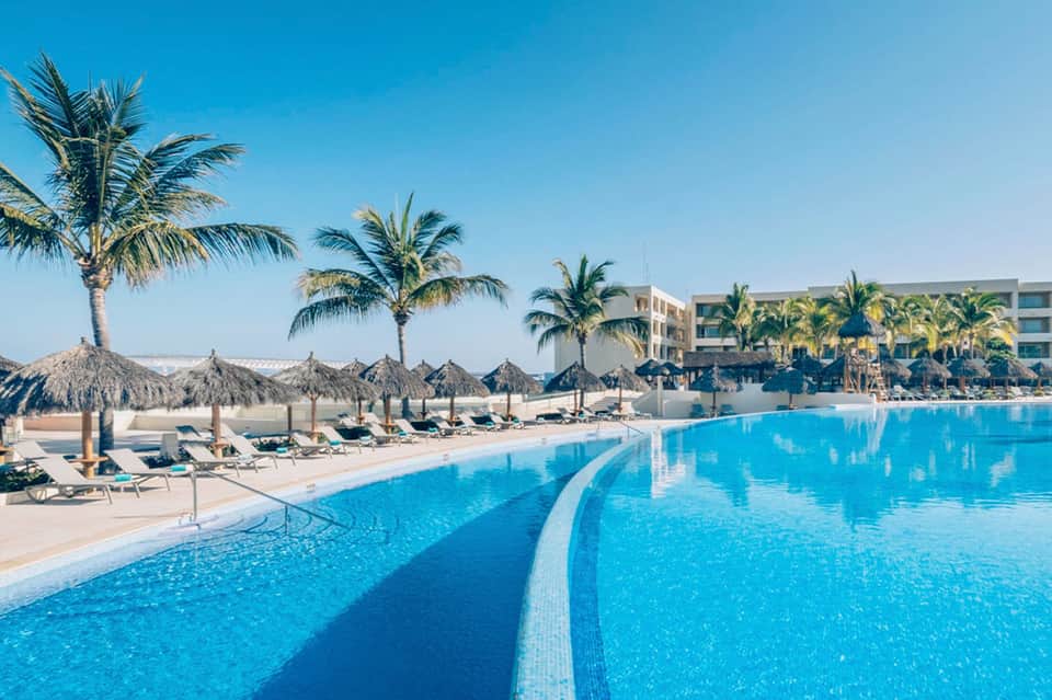 Large resort pool with palm trees, umbrellas, loungers, and beachfront hotel building