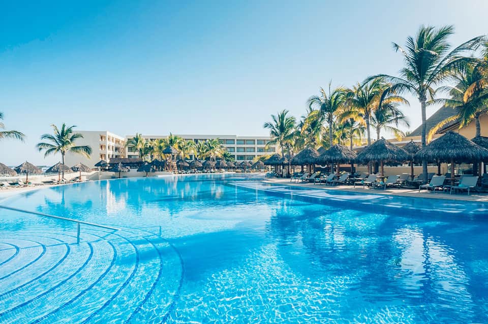 Large resort pool with turquoise water, tiki umbrellas, lounge chairs, and palm-lined beachfront