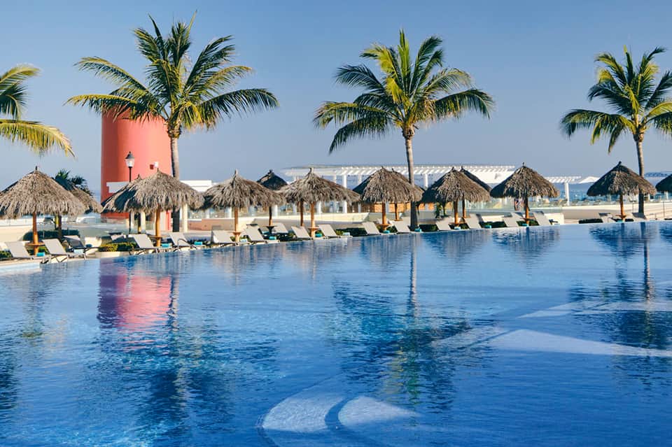 Modern resort pool with thatched cabanas, palm trees, and pink tower building