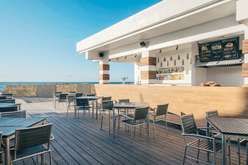 Modern beachfront bar with wooden counter and seating overlooking ocean under clear sky