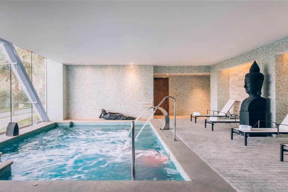 Modern indoor pool with turquoise water, Buddha statue, lounge chairs, and stone walls
