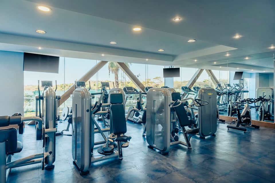Contemporary gym with floor-to-ceiling windows, geometric ceiling beams, and rows of cardio equipment