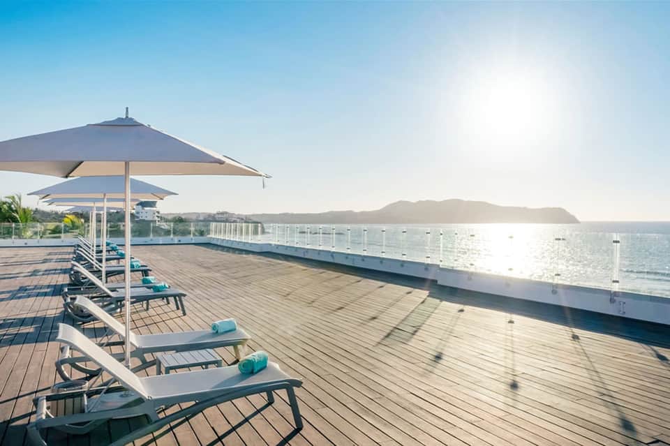 Oceanfront deck with white loungers and umbrellas overlooking a calm bay with distant mountains