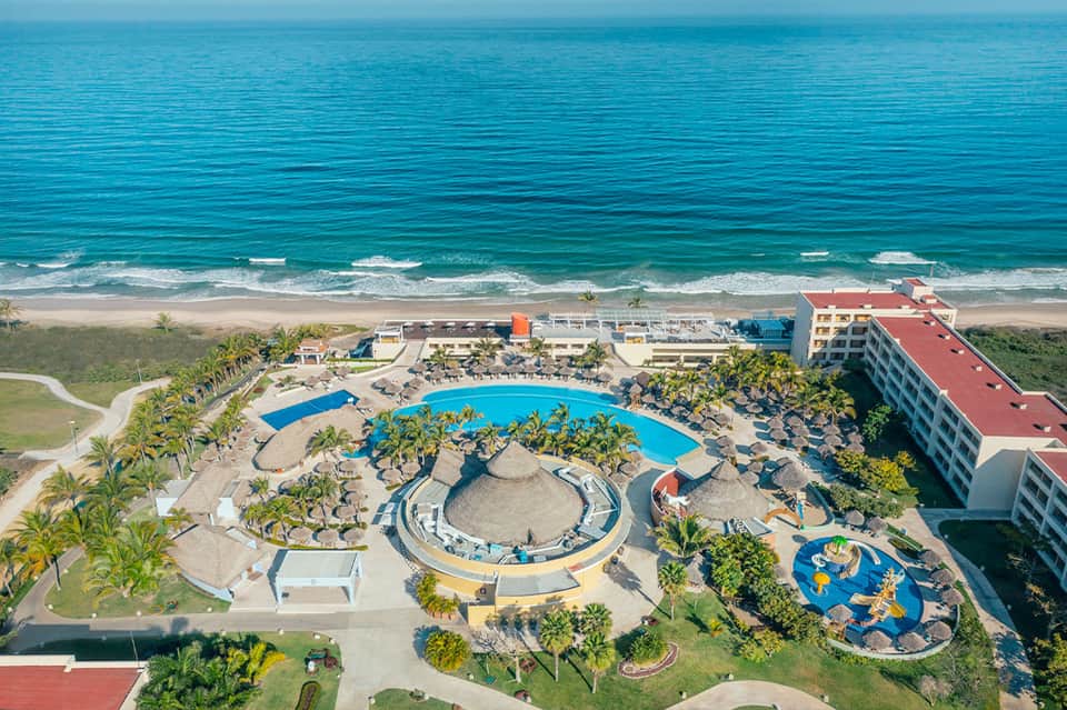 Aerial view of beachfront resort with large pool, palm trees, buildings, and sandy beach
