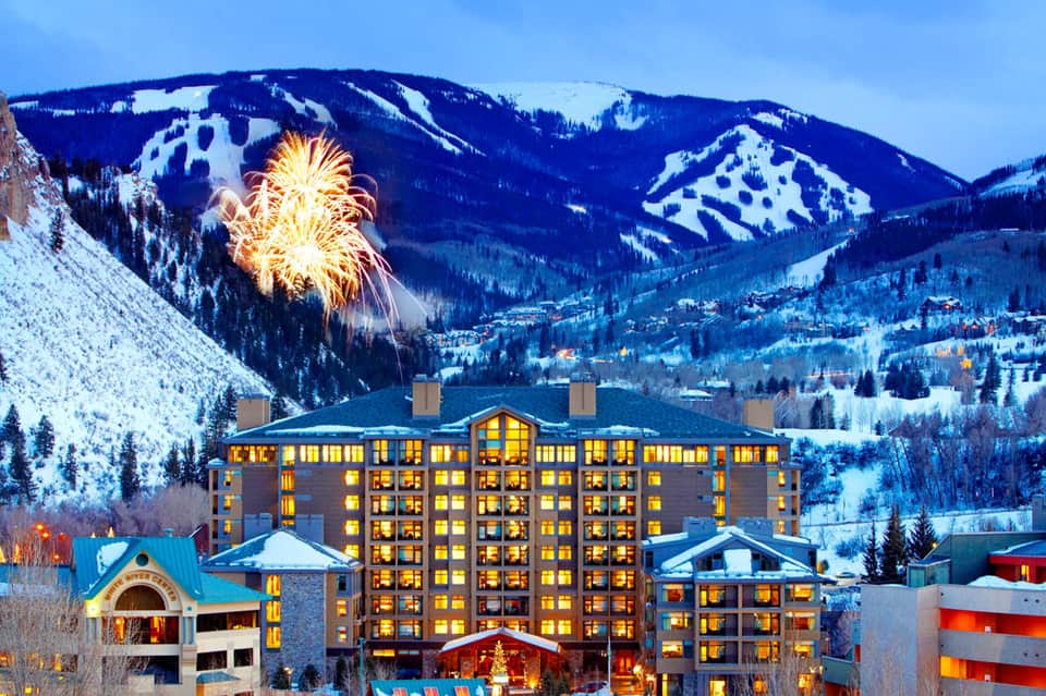 Luxury resort hotel with illuminated windows set against snow-covered mountains with fireworks display overhead