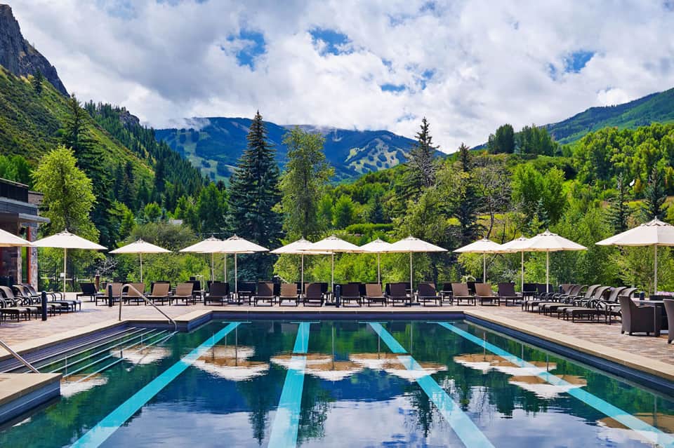 Outdoor lap pool with lane markers, lounge chairs, white umbrellas, and forested mountain views