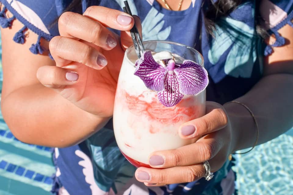 Hands holding tropical cocktail with purple orchid garnish by poolside
